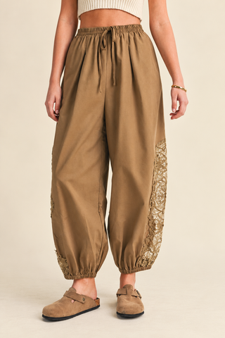 Women's camel balloon pants with floral lace inset detailing, elastic drawstring waistband, relaxed fit, and gathered ankle cuffs, styled with tan clogs.